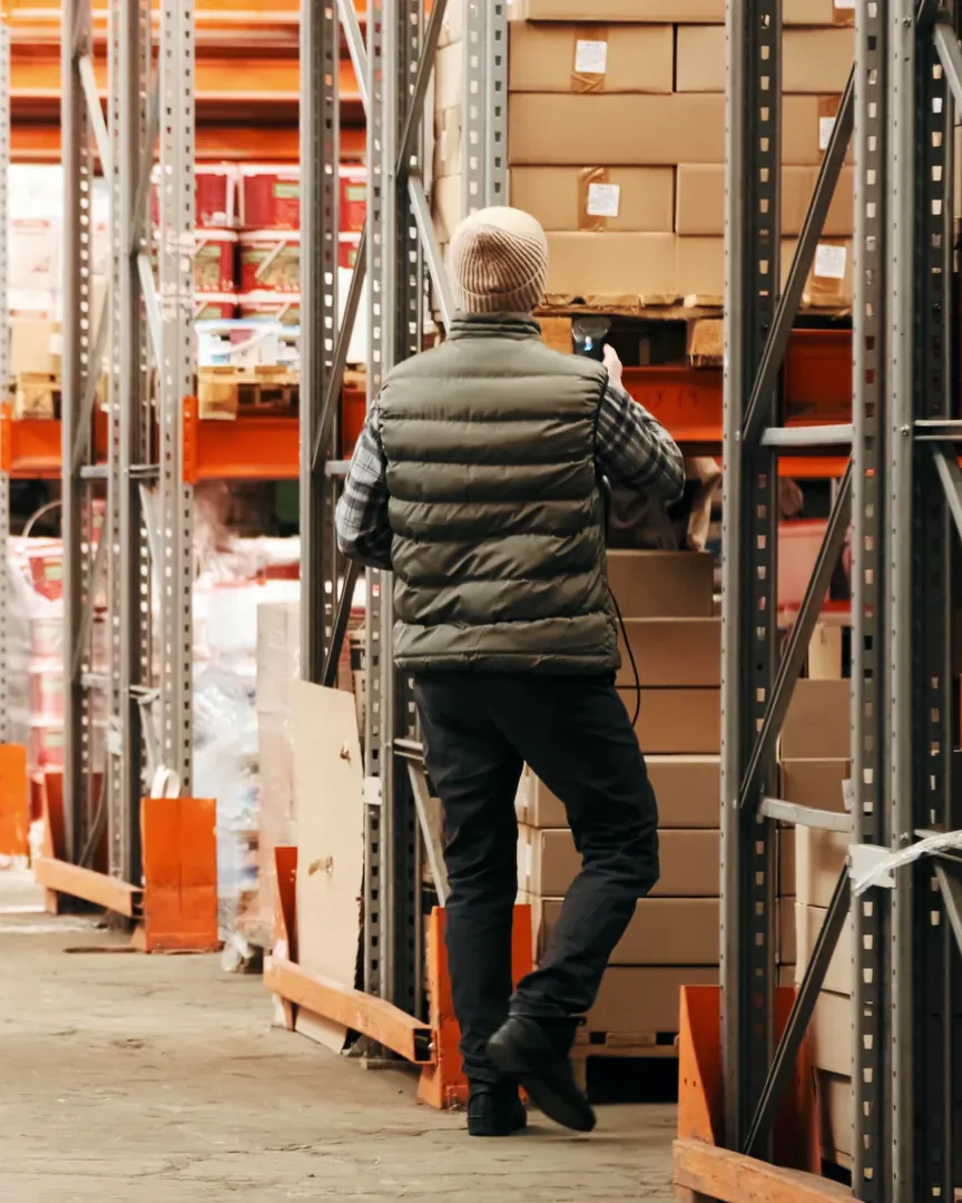 A person seen from behind in a padded vest and a woollen hat stands at a rack in a long warehouse aisle, reaching to a shelf of cartons between gray and orange uprights, a still from a stock film