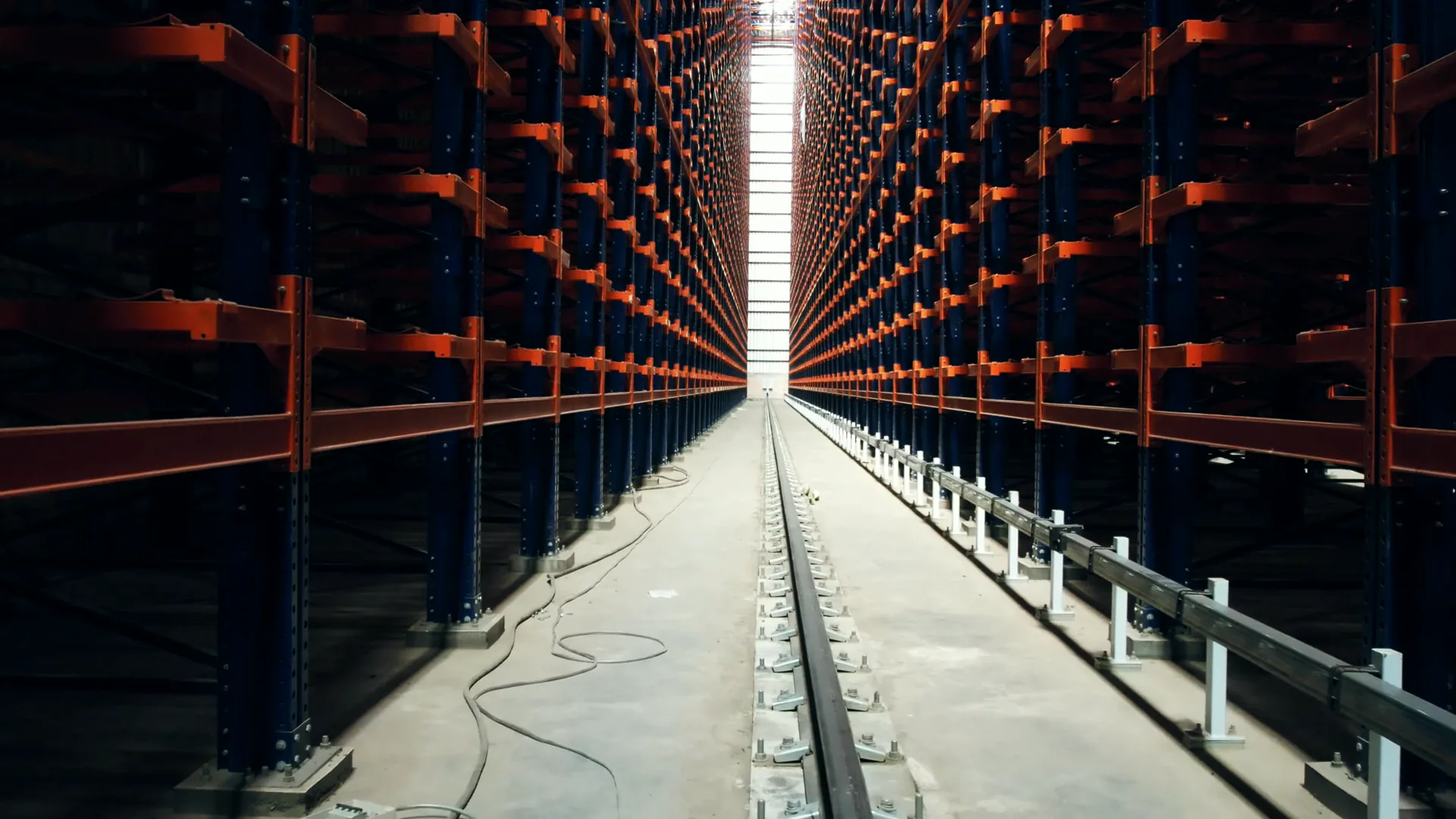 Looking down a narrow aisle of an automated high-bay store: blue uprights and orange beams on both sides, a shuttle rail in the floor and a slot of daylight at the far end, the first frame of a looping stock film
