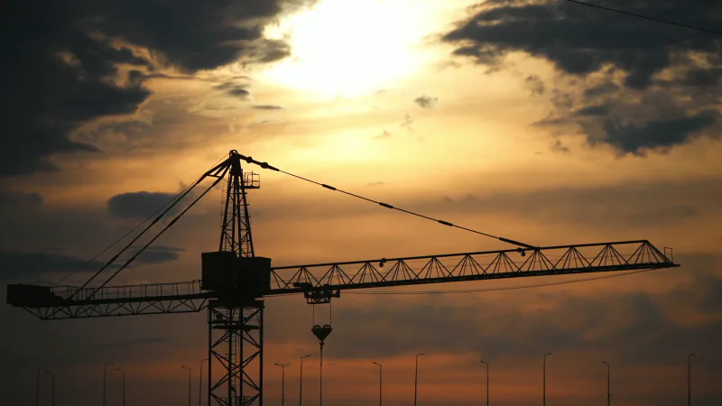 A single tower crane in silhouette with a hoist block hanging from its jib, a white sun breaking through dark clouds above, stock footage