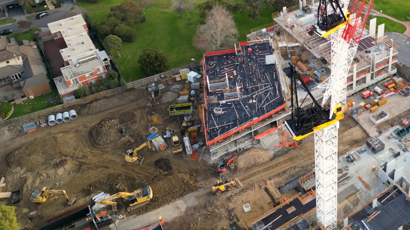 A building site seen obliquely from a drone: a slab under black sheeting beside a white tower crane, three excavators and a telehandler working a muddy platform, a few people a few pixels tall, stock footage with an illustration of event lines and boxes