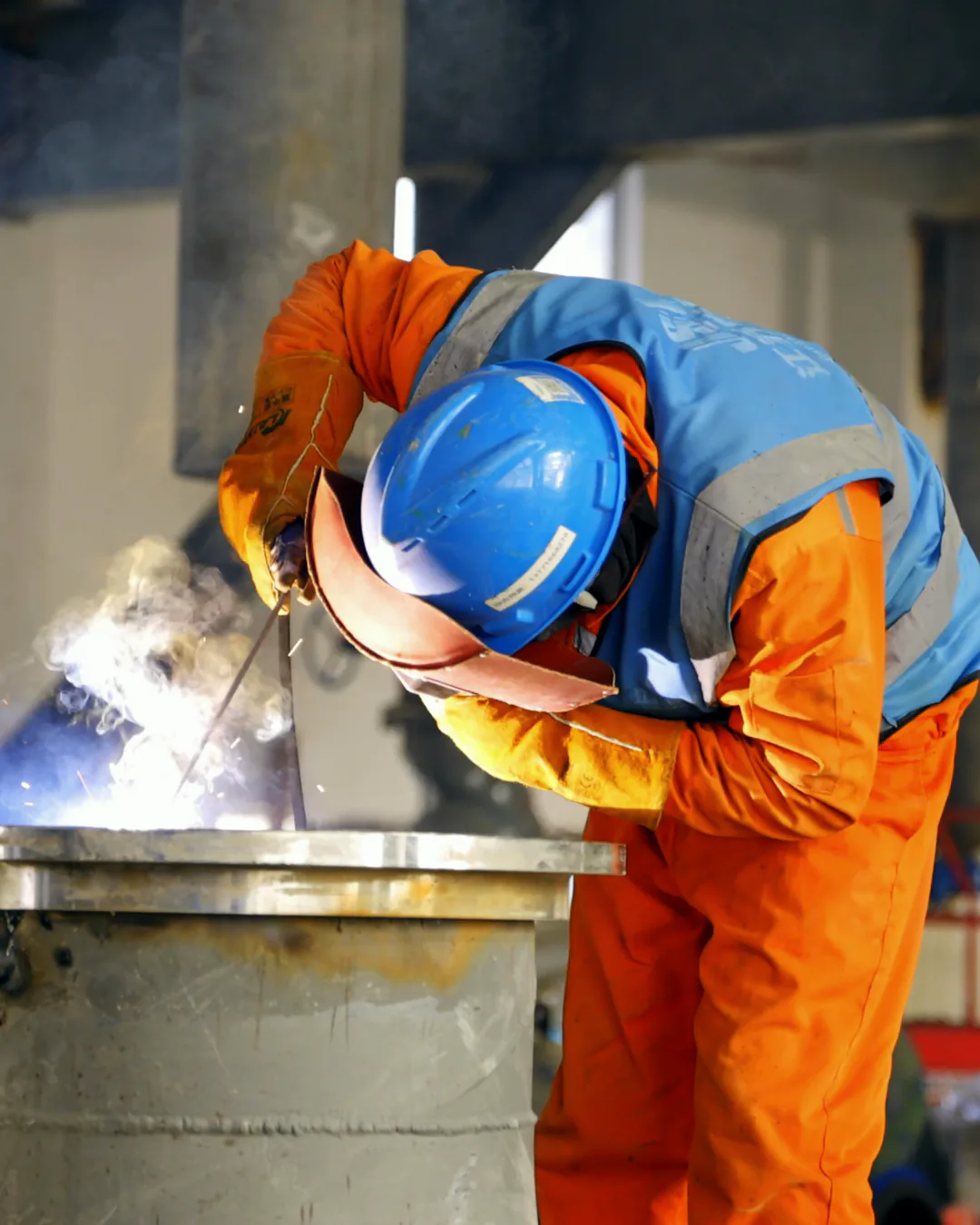A welder in an orange coverall and a blue vest bends over a steel drum, a bright arc at the lower left, a blue hard hat and a welding visor hiding his face, a still from a stock film