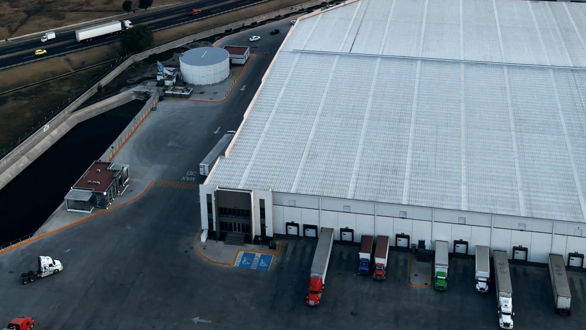Dusk over a distribution center seen from a drone: red, white and green trailers backed to a row of dock doors and a white tractor in the yard, the first frame of a looping stock film of four working sites: a dock face, an empty distribution hall, a steel rolling line and a rail yard