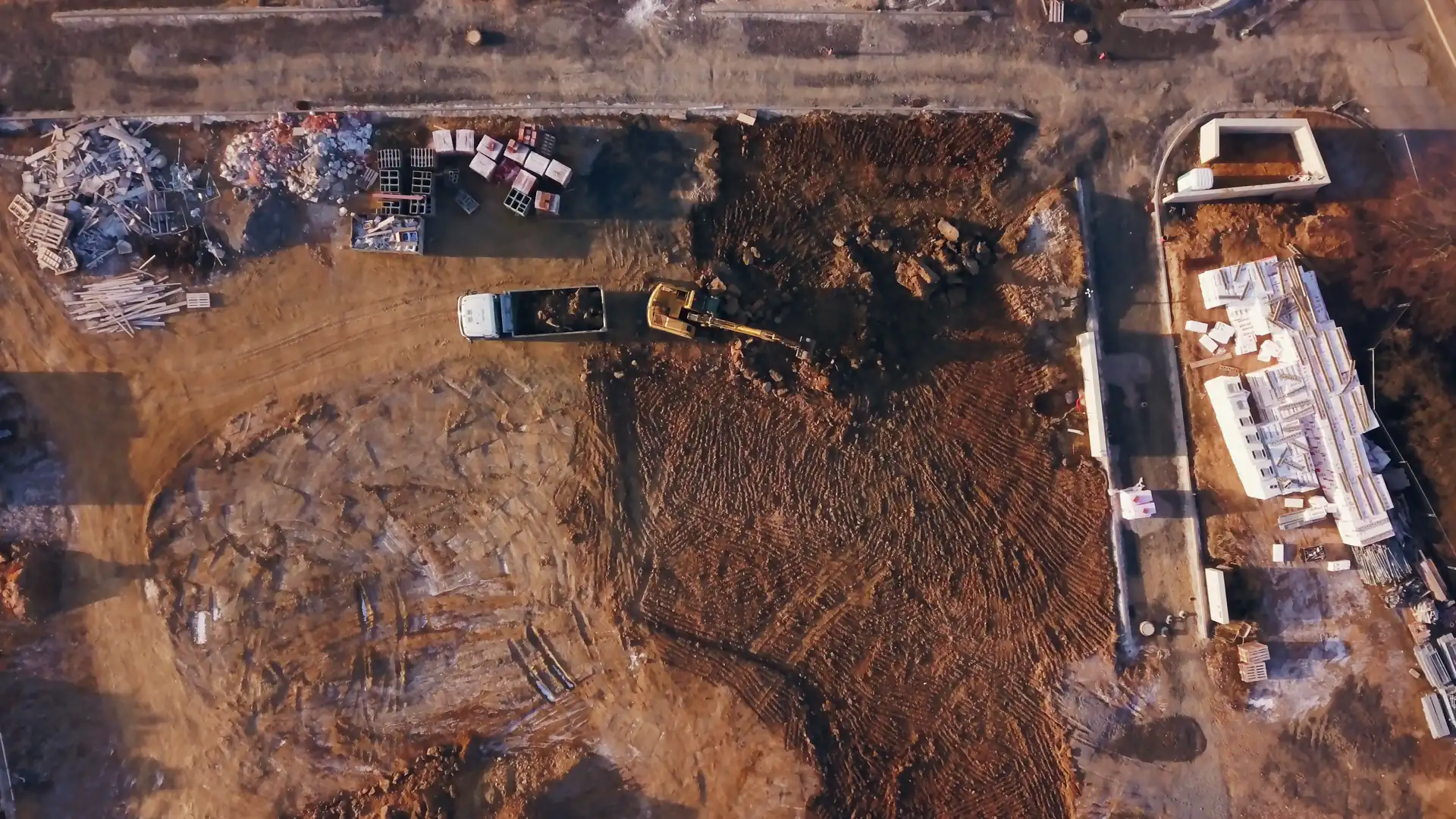 A building site seen from straight above in low golden light: a yellow excavator beside a white tipper truck loading it with soil, a churned plot, stacked white blocks and pallets, the first frame of a looping stock film, no people