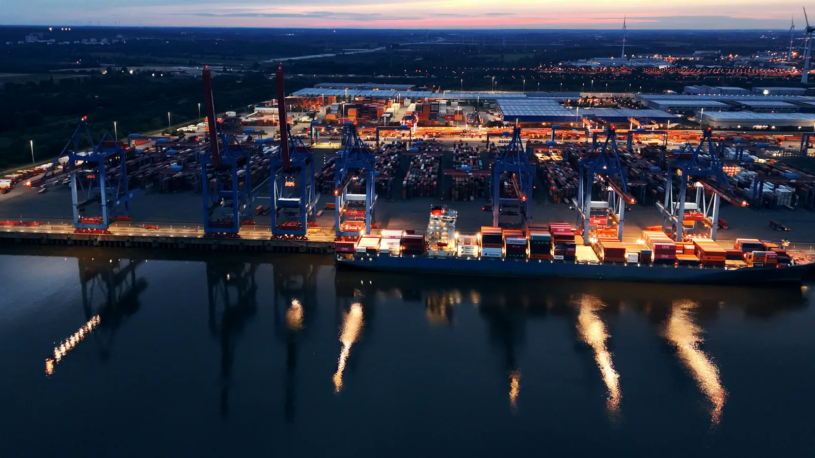 A container port at dusk seen from the air, lit gantry cranes and a loaded ship at the quay, the dark water below reflecting the lights, stock footage still