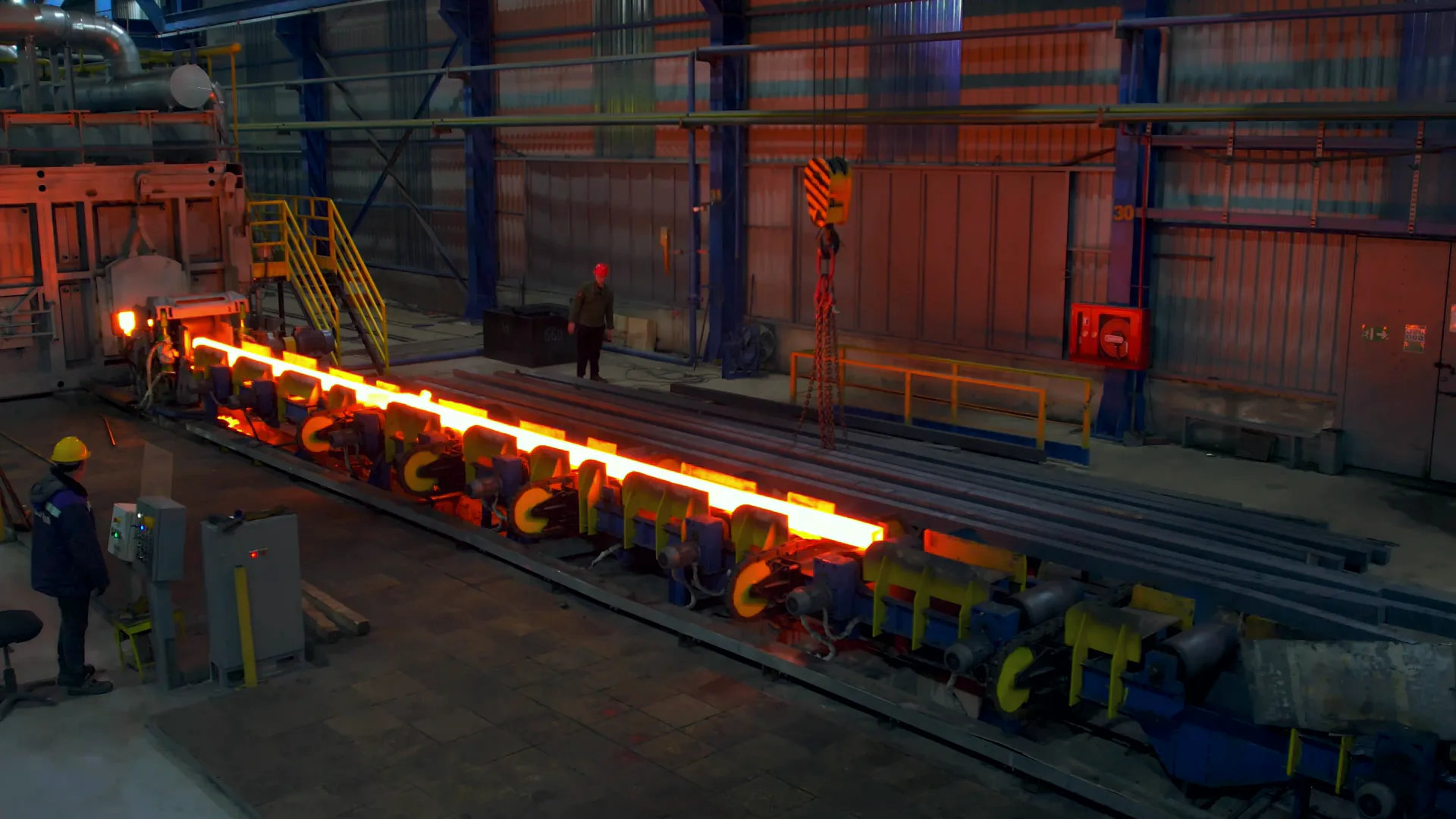 A steel rolling line in a hall at dusk: a white-hot bar runs along yellow roller stands from the furnace at the left, a worker in a hard hat at the left edge and another walking behind, a hoist hook and a red cabinet on the blue steelwork, the first frame of a looping stock film